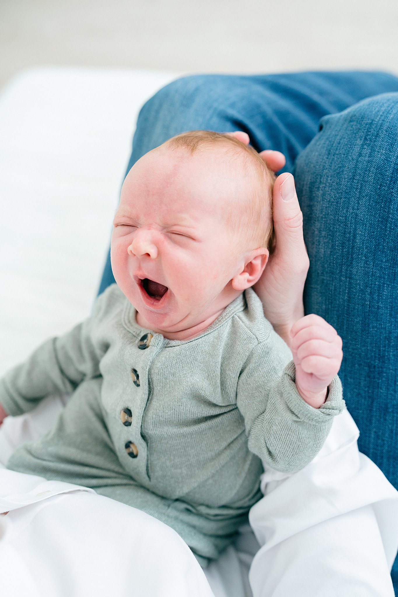 A newborn baby yawns while laying in a green onesie in mom's lap