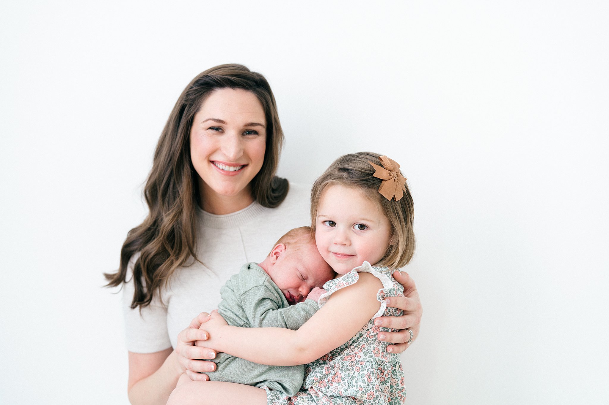 A newborn sleeps in big sisters lap with mom hugging them both in a studio after visiting atlanta baby boutiques