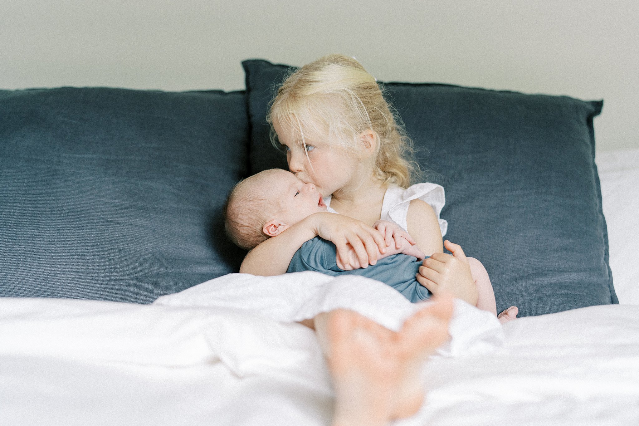 A toddler girl in a white dress sits on a bed kissing her newborn sibling in her lap