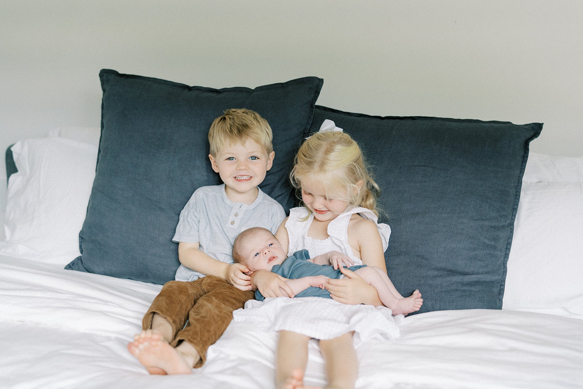 Happy toddler brother and sister sit on a bed holding their newborn sibling after visiting atlanta lactation consultants