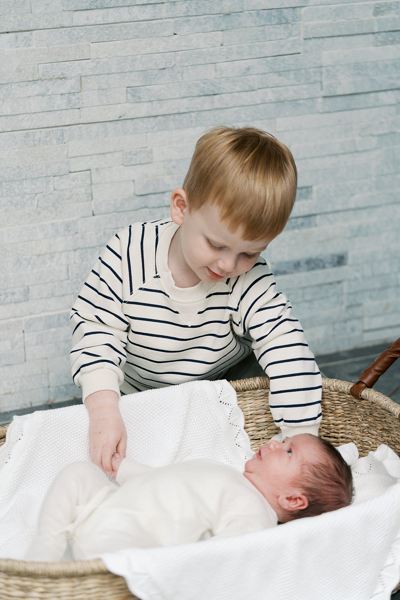 A toddler boy in a stripe shirt reaches into a basket his newborn sibling looks down onto