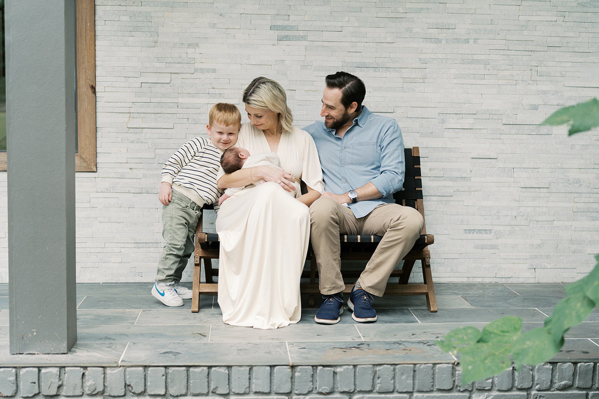 A happy mom and dad sit on a porch bench with their toddler son smiling at newborn in mom's lap after visiting atlanta pediatricians