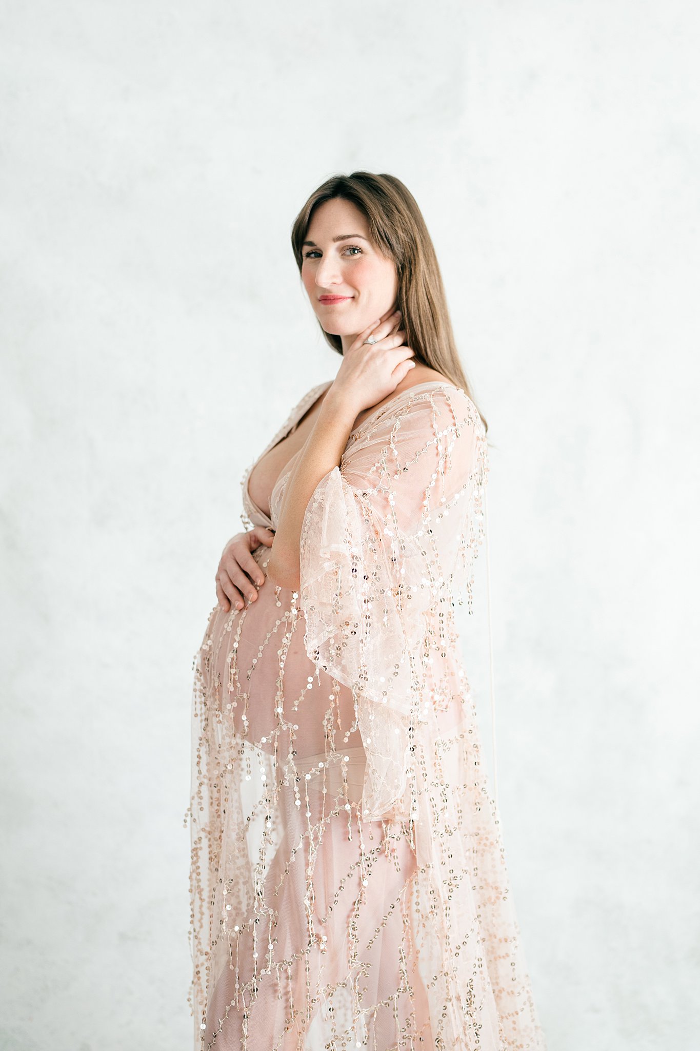 A happy mom to be rests a hand on her bump while standing in a studio in a sheer pink gold maternity gown