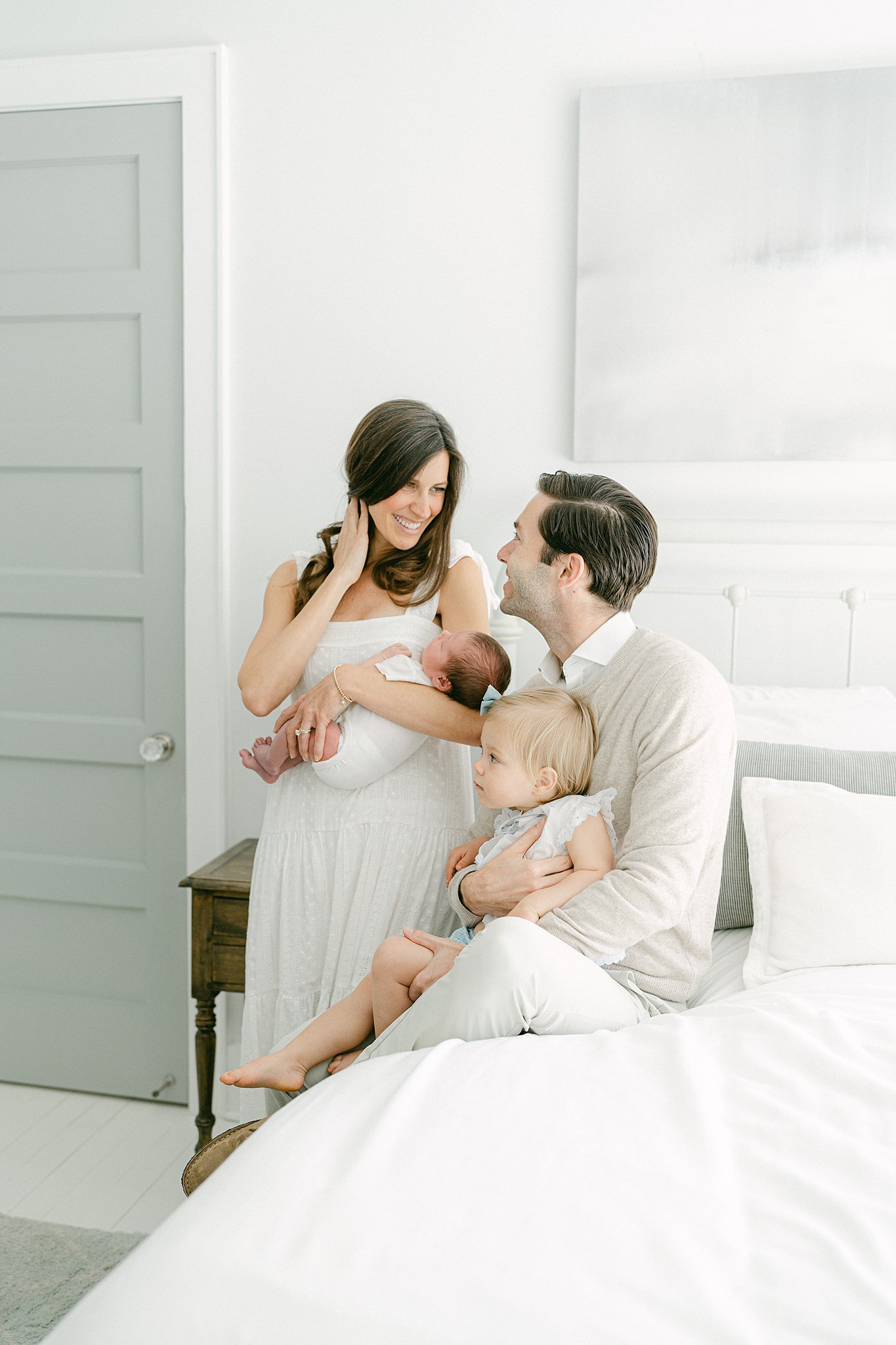 A happy mom and dad smile at each other while holding their newborn and toddler daughter in their bedroom after leaving birthing centers in Atlanta