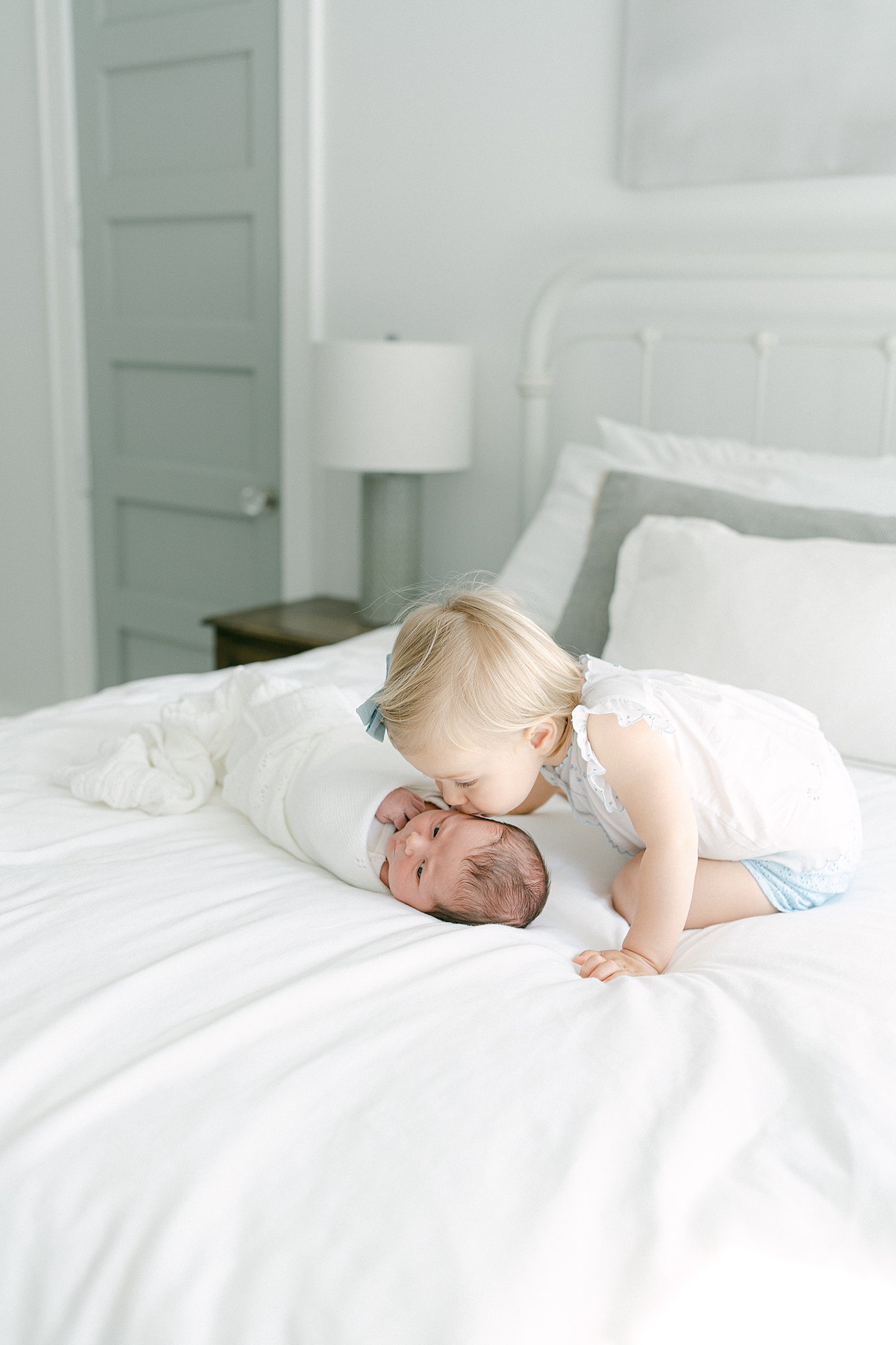 A toddler on a bed leans over to kiss her newborn baby sibling in a white swaddle after leaving birthing centers in Atlanta