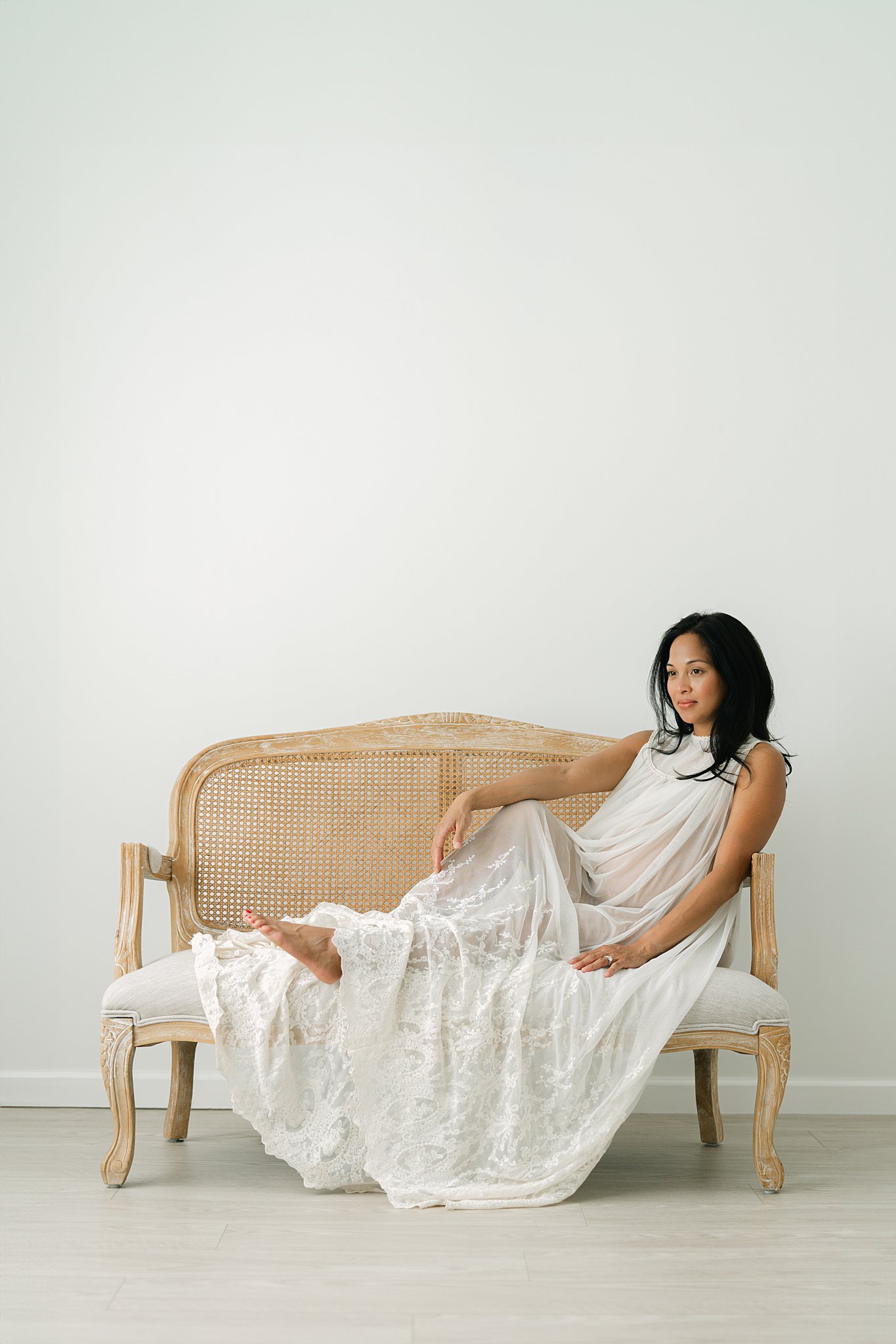 A pregnant woman sits across a bench in a studio in a long white gown