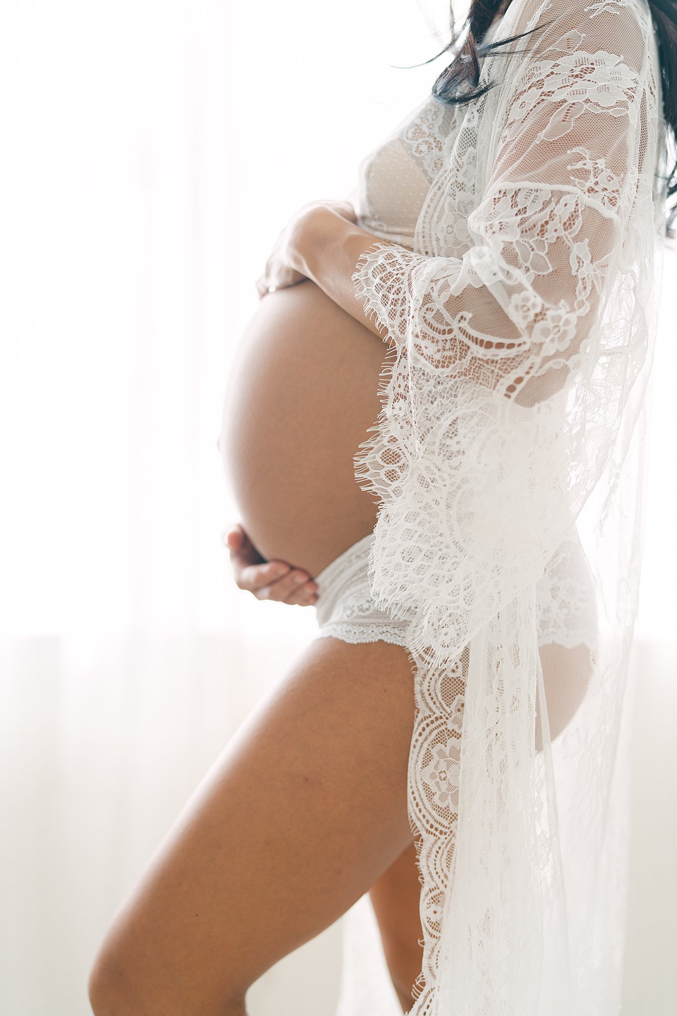 A mom to be stands in a studio in white lace lingerie in a window holding her bump after a prenatal massage in Atlanta